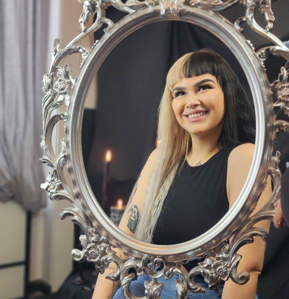 A woman is smiling at home in front of an ornate mirror.