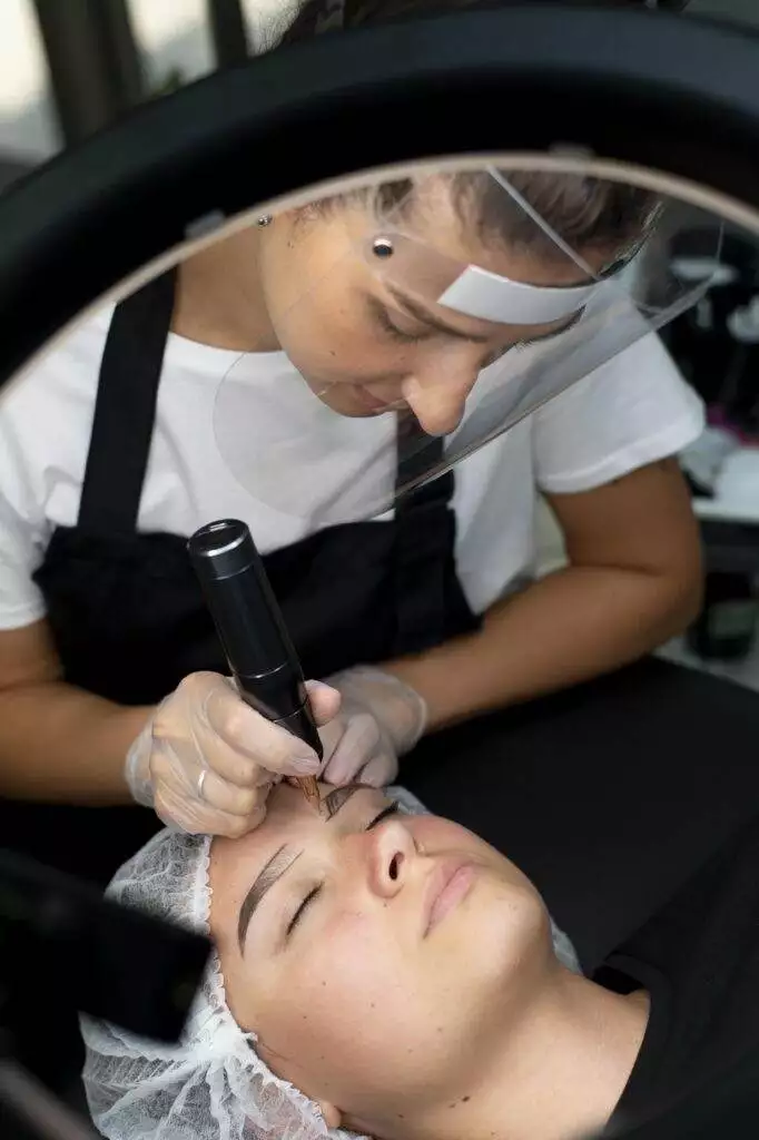 Aesthetician using a handheld device to perform eyebrow tattooing on a relaxed female client lying down, both under a ring light.