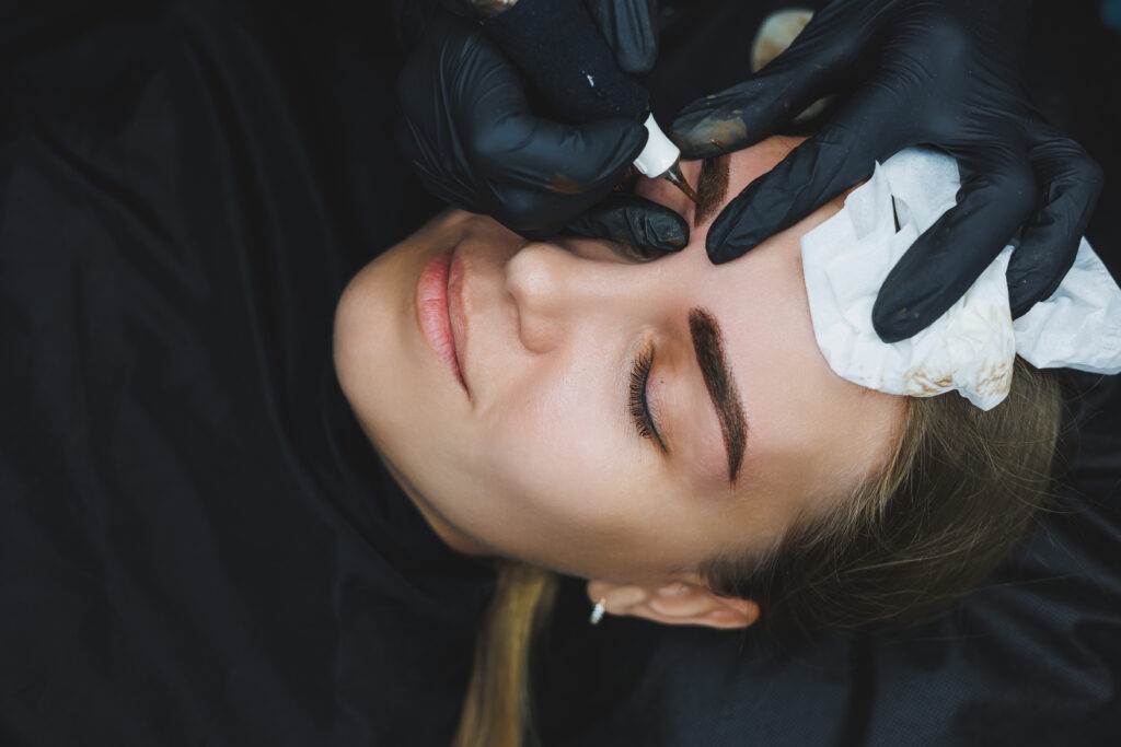 A woman receiving an eyebrow tattoo at a salon, with a beautician wearing gloves applying Dark Heart Ink to her eyebrows.