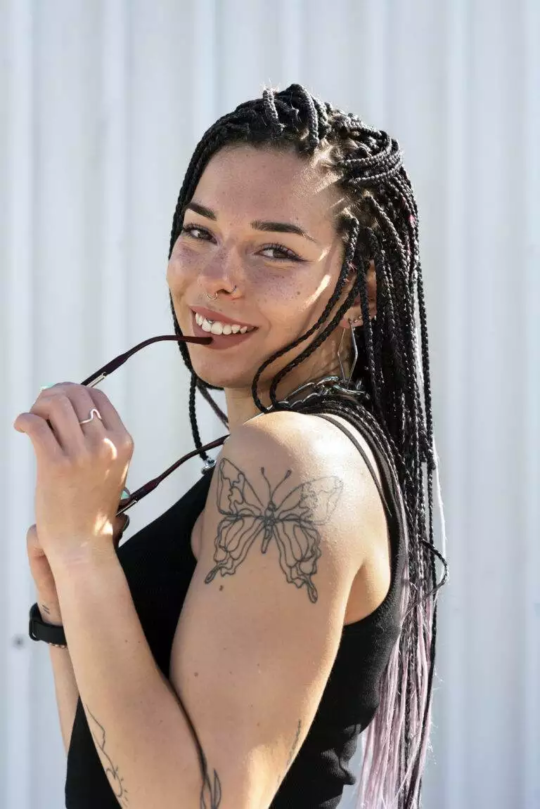 A young woman with braided hair and cosmetic freckle tattooing smiles, holding sunglasses, against a corrugated metal background.