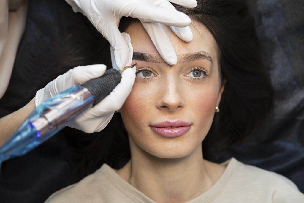 A woman receiving a cosmetic tattoo procedure at Dark Heart Ink, with a specialist holding a microneedling device near her forehead.