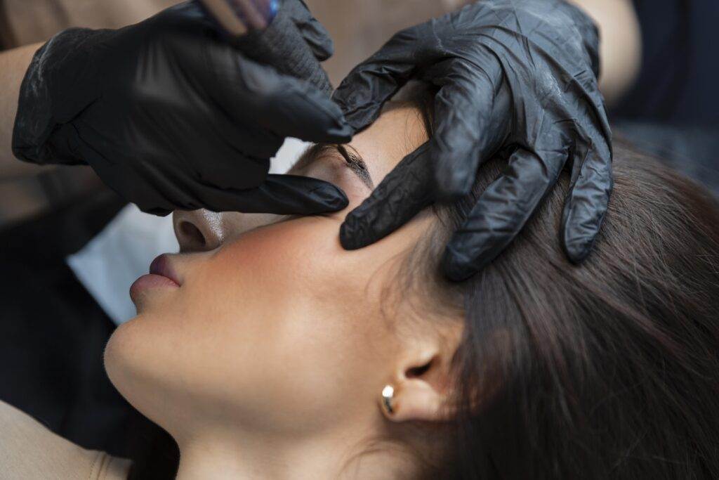 A professional beautician wearing black gloves maintains a woman's eyebrow tattoos at Dark Heart Ink in a close-up view.