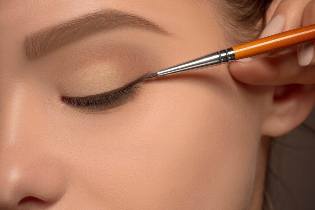 Close-up of a hand applying black eyeliner with a brush to a person’s closed eyelid, transforming the look from classic to dramatic.