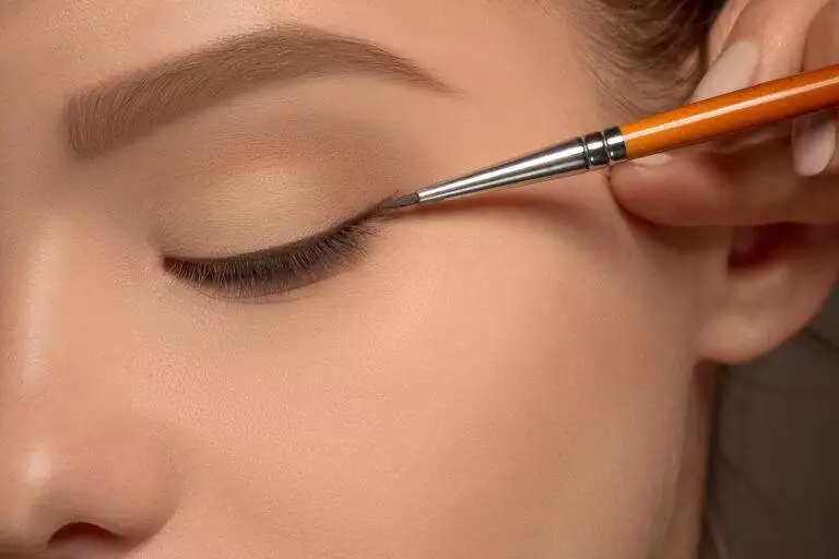 Close-up of a hand applying black eyeliner with a brush to a person’s closed eyelid, transforming the look from classic to dramatic.