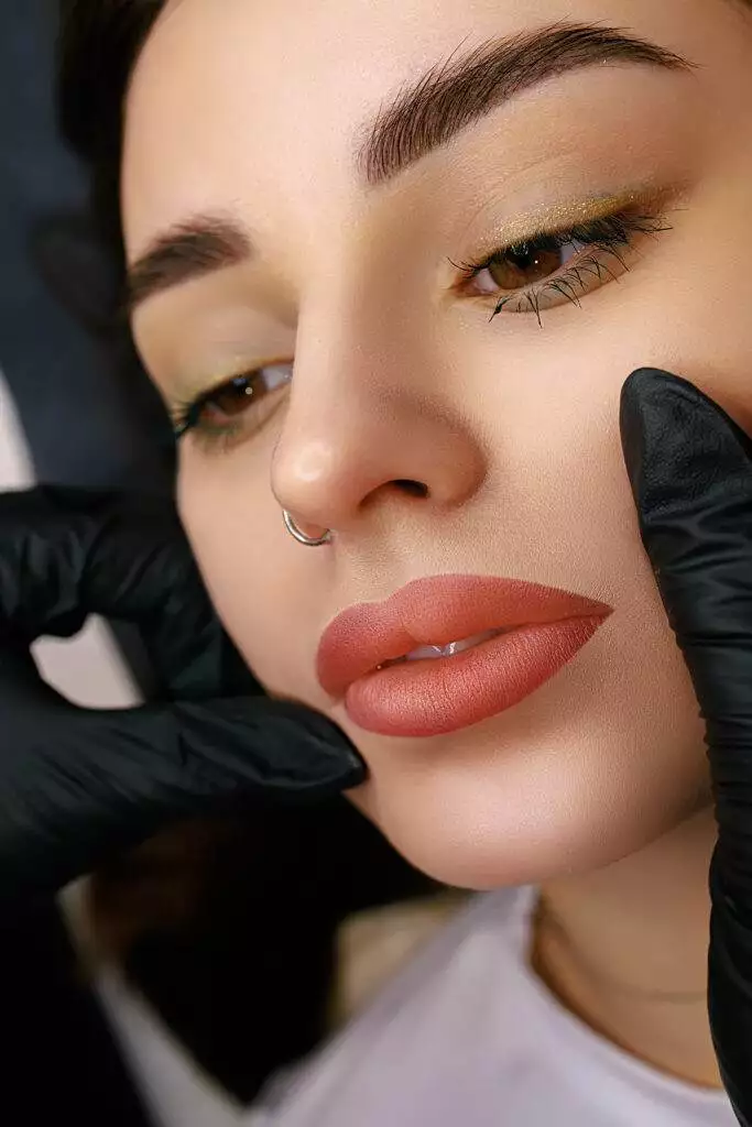 Close-up of a woman's face with bold makeup and a nose ring, capturing the benefits of lip blushing. She is being gently touched on the jaw by hands wearing black gloves.