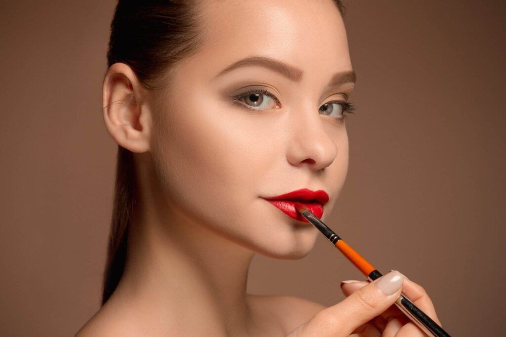 A person with fair skin applies red lipstick with a brush, looking directly at the camera against a brown background, showcasing the benefits of lip blushing for San Diego residents seeking long-lasting color.