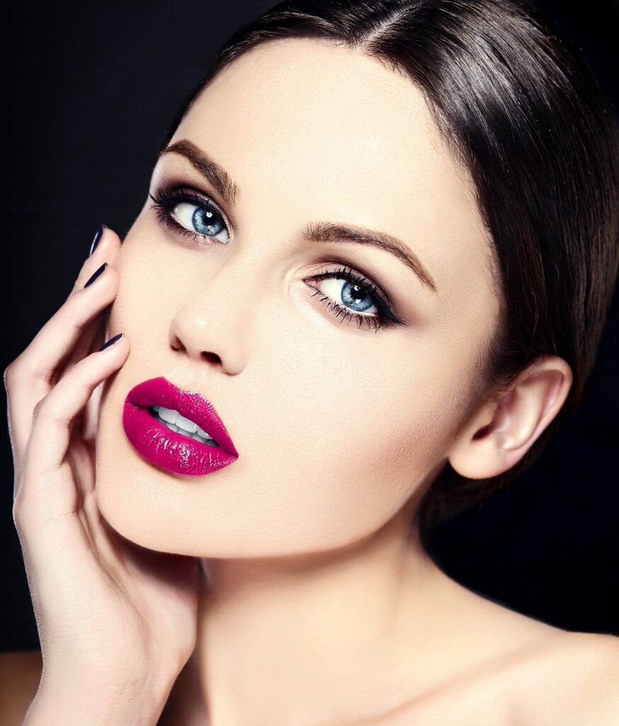 A woman with striking blue eyes, bold pink lipstick, and dramatic eye makeup—perfect for busy women opting for permanent makeup in San Diego—lightly touching her cheek, set against a dark background.