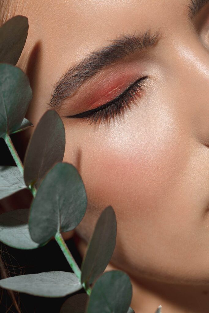 Close-up of a person's face with closed eyes and pink eye shadow, framed by green eucalyptus leaves, showcasing the elegance of permanent eyeliner.