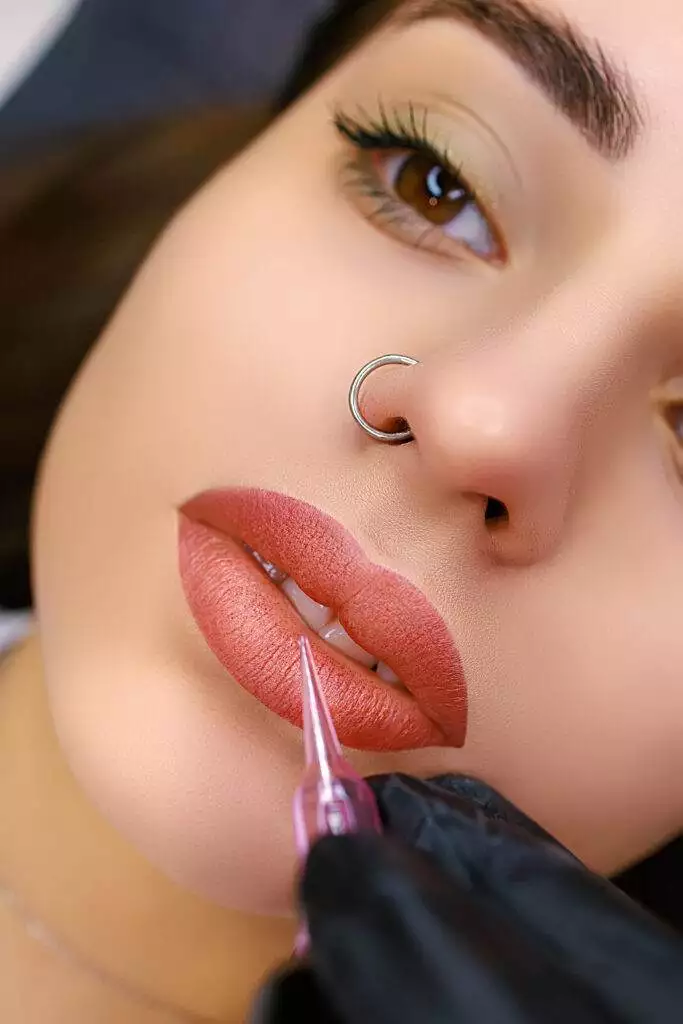 A close-up of a person with a nose ring receiving a cosmetic lip blushing procedure with a pink tool held by a gloved hand, showcasing the benefits for San Diego residents.