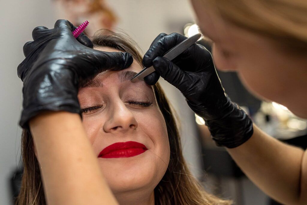 A woman with red lipstick receives eyebrow grooming from a person wearing black gloves, using tweezers and a brush, ensuring flawless results while at Dark Heart Ink.