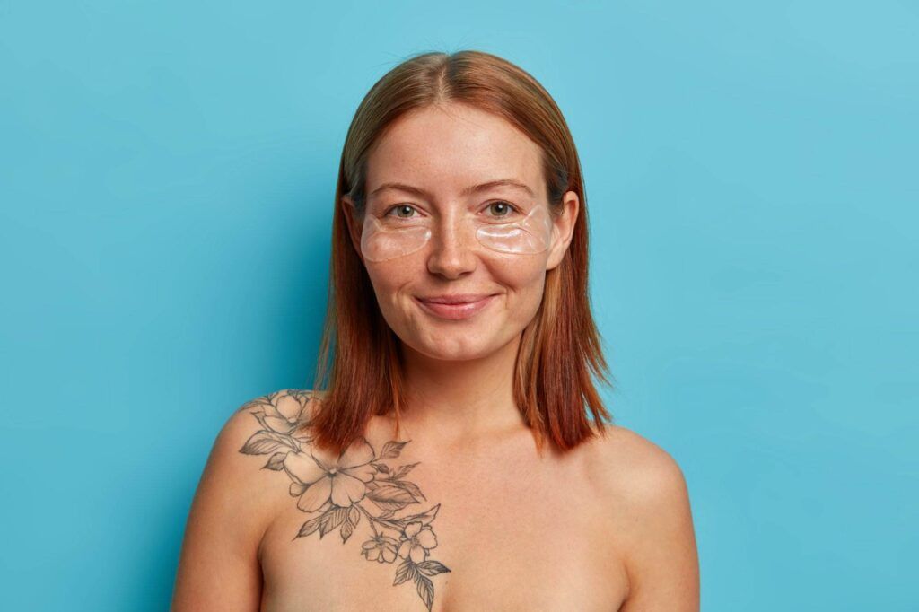 A woman with shoulder-length red hair stands against a blue background, wearing under-eye masks and displaying a floral tattoo from Dark Heart Ink on her left shoulder. Her expression exudes a sense of inner beauty.