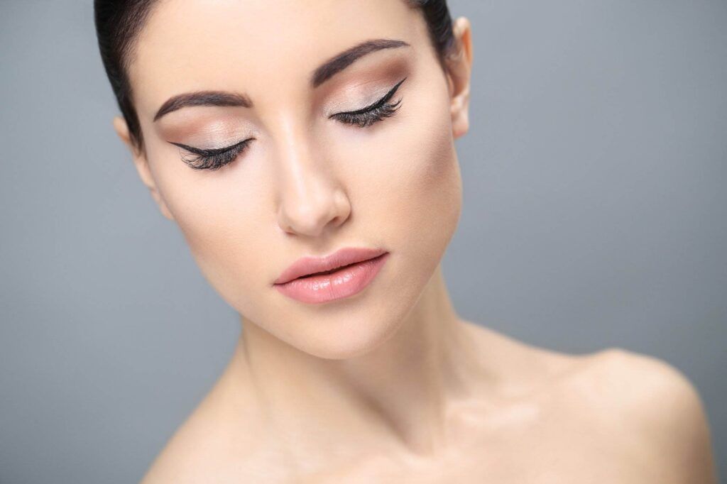 Close-up of a woman's face with closed eyes, featuring well-defined eyebrows, winged eyeliner in one of the popular Permanent Eyeliner Styles, and nude lipstick. The background is a neutral gray.