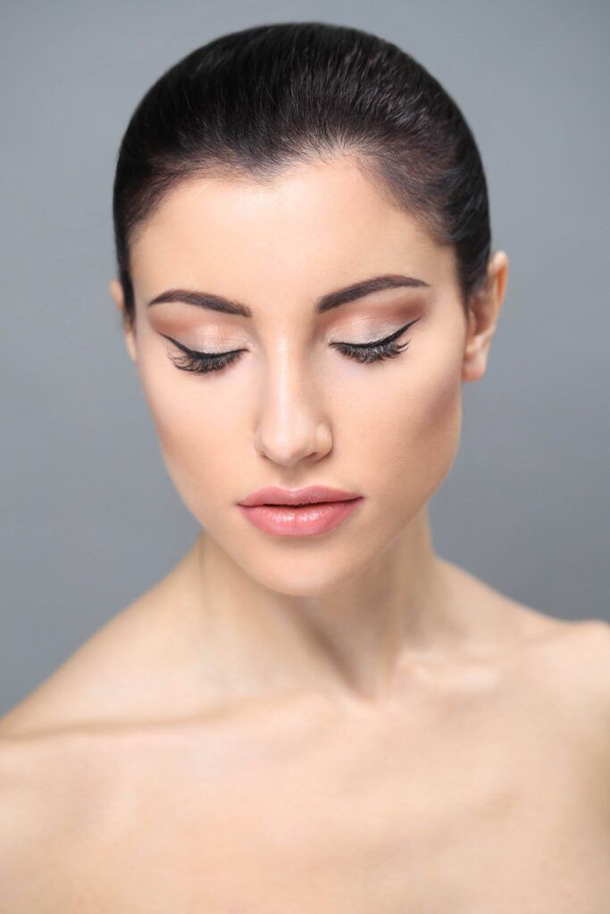 A portrait of a woman with smooth skin and dark, slicked-back hair. She has winged eyeliner, embracing one of the Permanent Eyeliner Styles from Dark Heart Ink, and is looking downward with a neutral expression against a plain gray background.