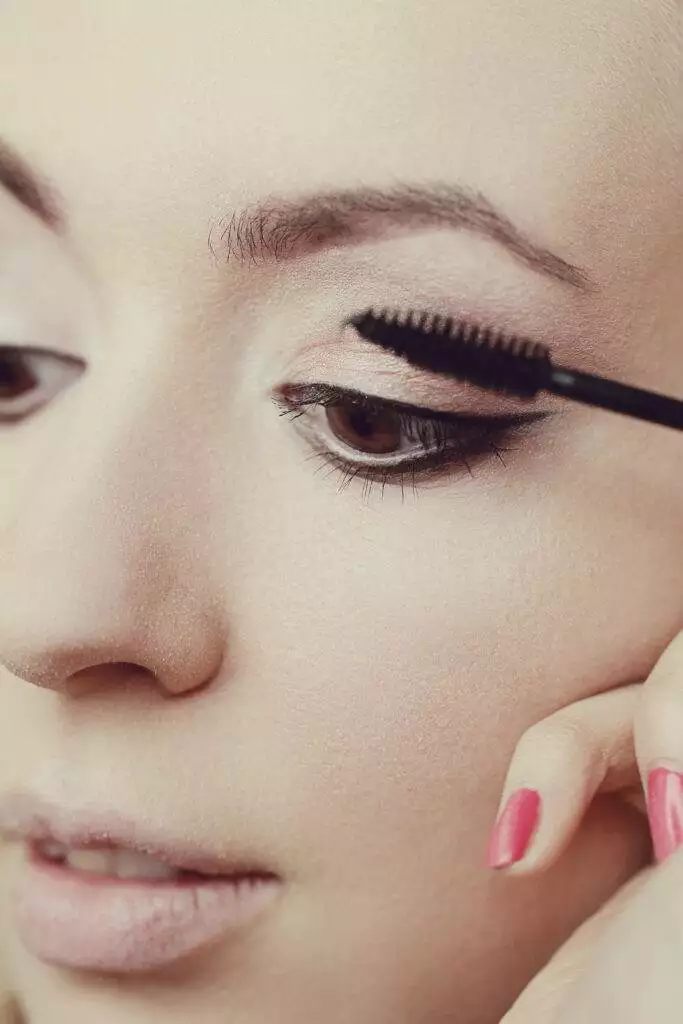 A close-up of a person applying mascara to their upper lashes. The person holds a mascara wand near their right eye and has pink painted nails, taking care to avoid any smudging while complementing their permanent eyeliner.