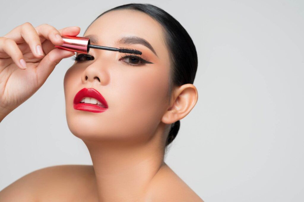 A person with styled hair applies black mascara to their upper eyelashes, showcasing bold eyeliner styles and wearing red lipstick. Their dark eyeliner, reminiscent of the craftsmanship at Dark Heart Ink, completes the striking look.