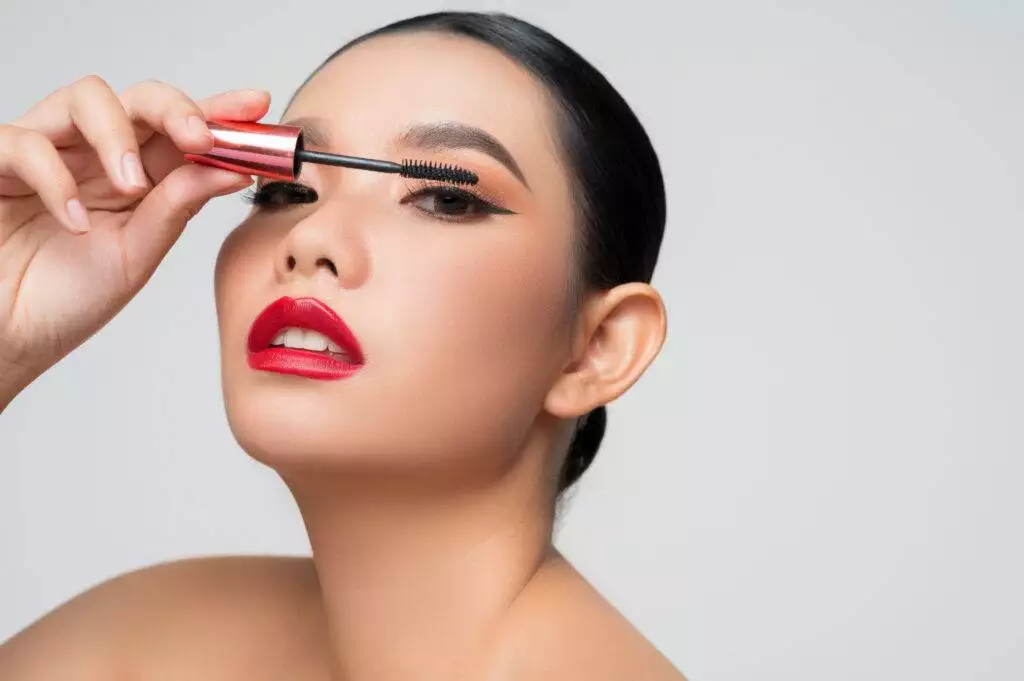 A person with styled hair applies black mascara to their upper eyelashes, showcasing bold eyeliner styles and wearing red lipstick. Their dark eyeliner, reminiscent of the craftsmanship at Dark Heart Ink, completes the striking look.