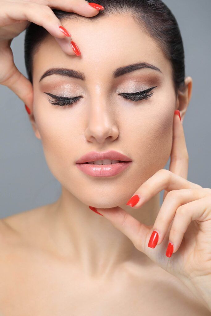 A woman with permanent winged eyeliner and striking red nail polish poses with eyes closed, hand touching her cheek and forehead against a neutral background.