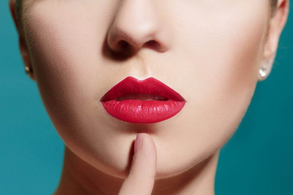 Close-up image of a person with red lipstick, lightly touching their lips with an index finger against a greenish-blue background, showcasing a perfect pout.