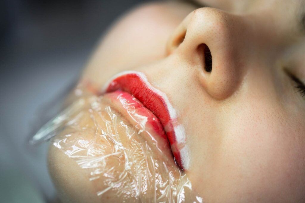 Close-up of a person's face with plastic wrap covering their lower face and lips, which appear to be outlined with a white and red substance, creating the illusion of a perfect pout.