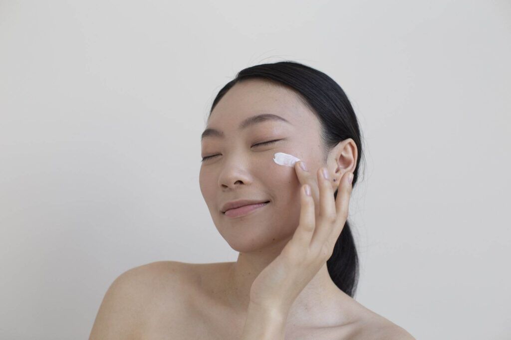 A person with closed eyes, natural brows complementing their serene expression, applies a cream to their cheek using their right hand.