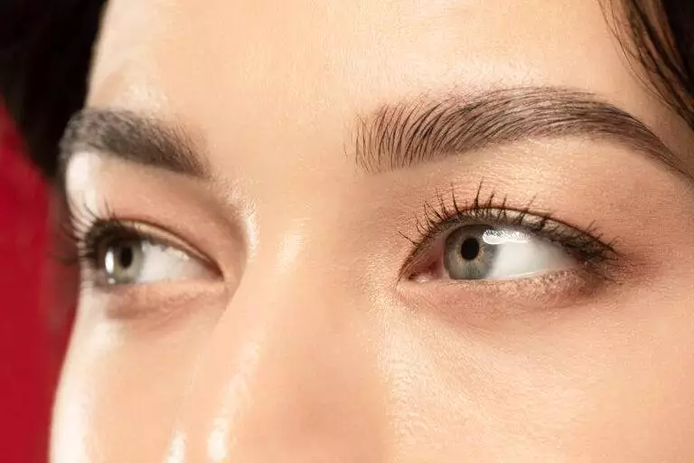 Close-up of a person's eyes looking slightly upward, showcasing well-defined eyebrows that could benefit from eyebrow tattooing. The image highlights long eyelashes and smooth skin against a solid red background.