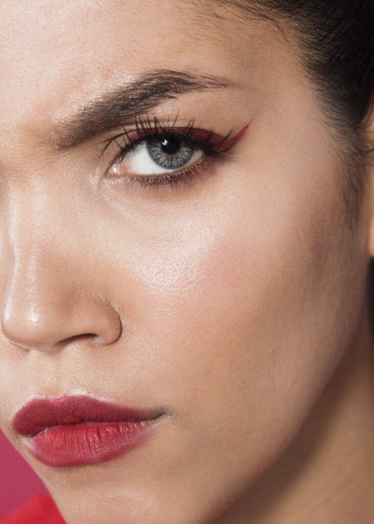 Close-up of a person with dramatic red makeup, including bold cosmetic winged eyeliner tattoos and lipstick, looking intensely at the camera, embodying effortless beauty.