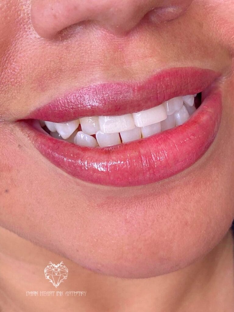 Close-up of a person's mouth with naturally enhanced lips in soft pink lipstick, showcasing well-aligned, white teeth. A small logo graces the bottom left corner, subtly blending style and elegance.