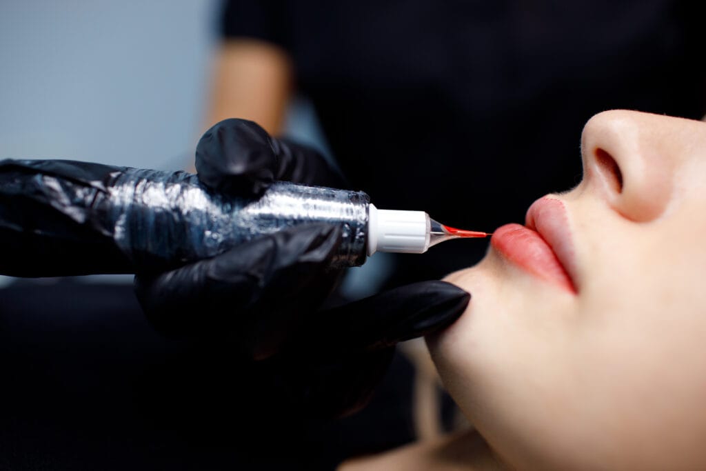 Close-up of a person's face receiving a cosmetic lip procedure, as a gloved hand expertly applies pigment using a tool. This lip blushing technique is popular in East Bay for achieving beautifully enhanced lips with a natural look.