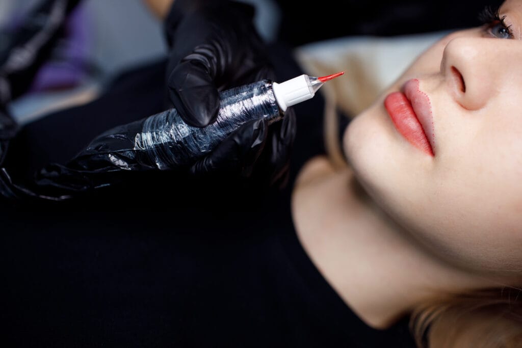 A person receives a lip tattoo procedure for naturally enhanced lips. The technician, wearing black gloves, skillfully uses a tattoo machine with red pigment near the lips to perfect the art of lip blushing.