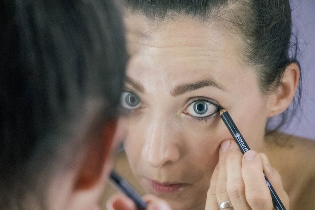A person applies eyeliner while looking in a mirror, focusing on their eye.