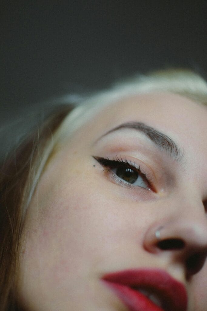 Close-up of a person's face angled upward, showing one eye with permanent eyeliner, defined eyebrow, nose piercing, and red lips—highlighting eyeliner benefits for a bold look in the East Bay.