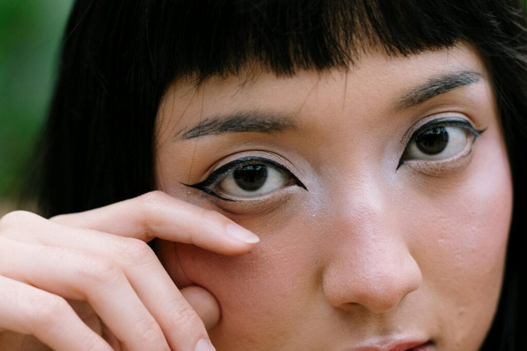 Close-up of a person with winged eyeliner and metallic eyeshadow touching their cheek with their fingers, showcasing the stunning benefits of permanent eyeliner available in East Bay.