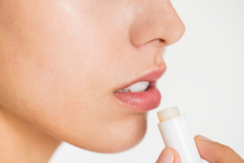 Close-up of a person applying lip balm to their lips with a white tube against a neutral background, highlighting essential Lip blushing aftercare for those living in the East Bay climate.